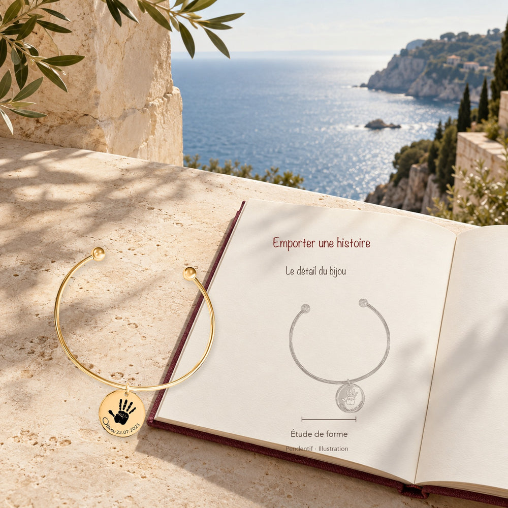 Bracelet jonc Empreinte de Main Réelle Gravée en finition dorée dans un décor méditerranéen, avec dessin du produit sur un carnet.