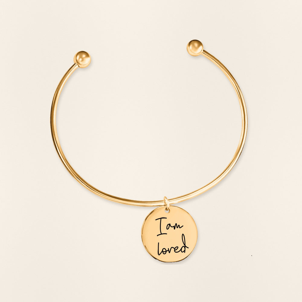 Bracelet jonc Médaille Affirmations Personnalisée en finition dorée, exemple de gravure sur fond ivoire texturé.