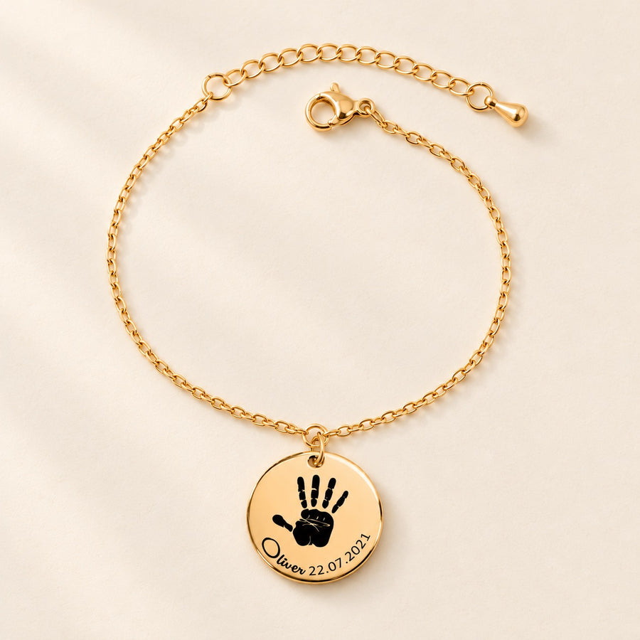 Bracelet chaîne doré gravé d’une empreinte de main et du prénom Oliver sur fond ivoire.