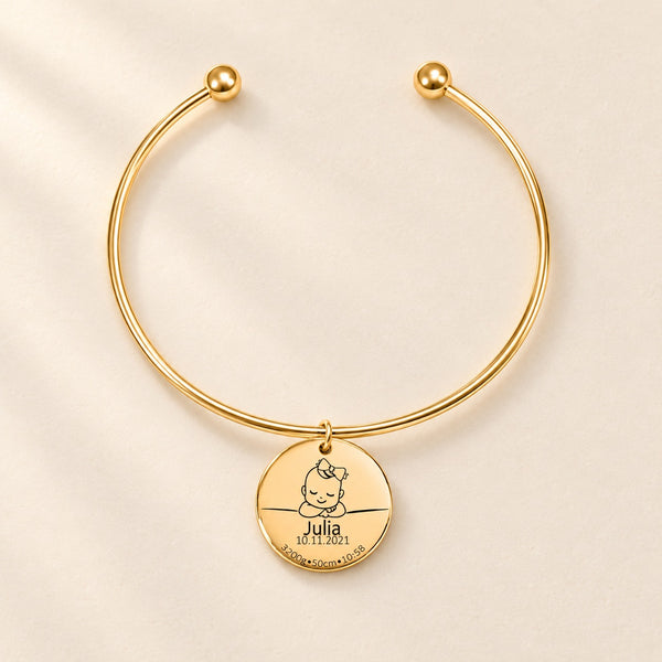 Bracelet jonc doré avec médaille de naissance gravée Julia sur fond ivoire.