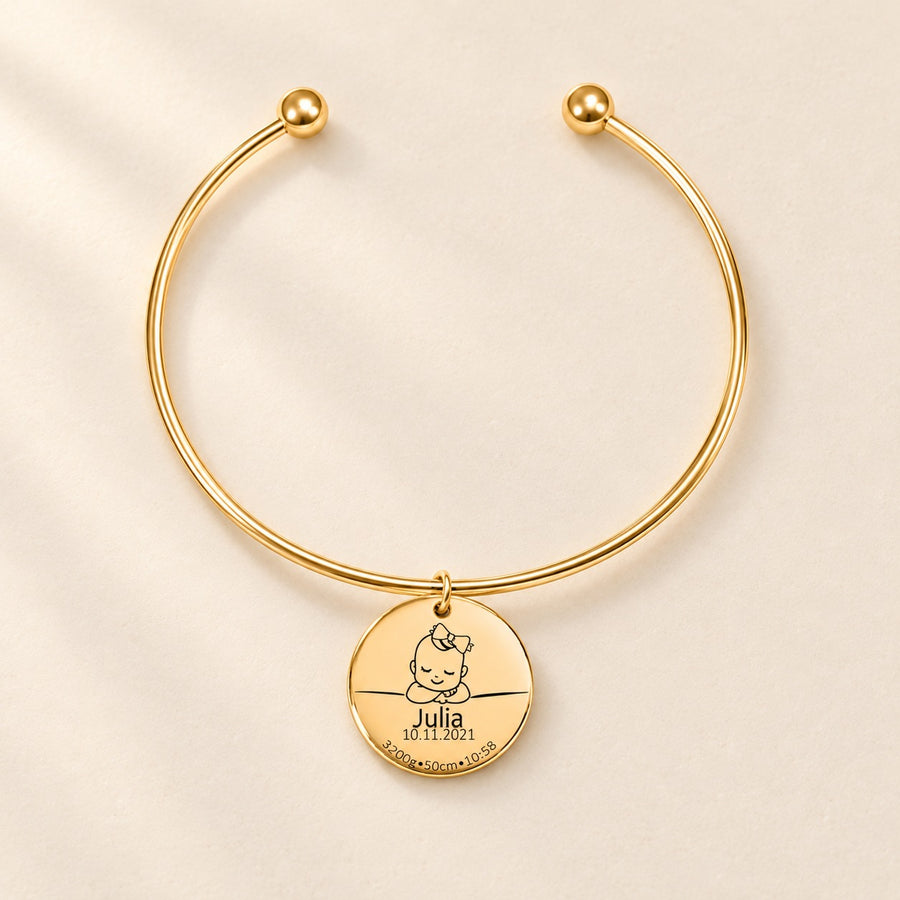 Bracelet jonc doré avec médaille de naissance gravée Julia sur fond ivoire.