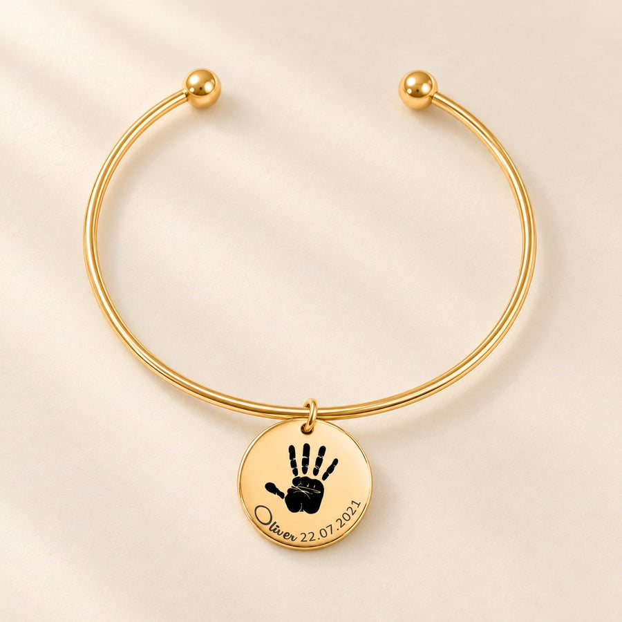 Bracelet jonc doré gravé d’une empreinte de main et du prénom Oliver sur fond ivoire.