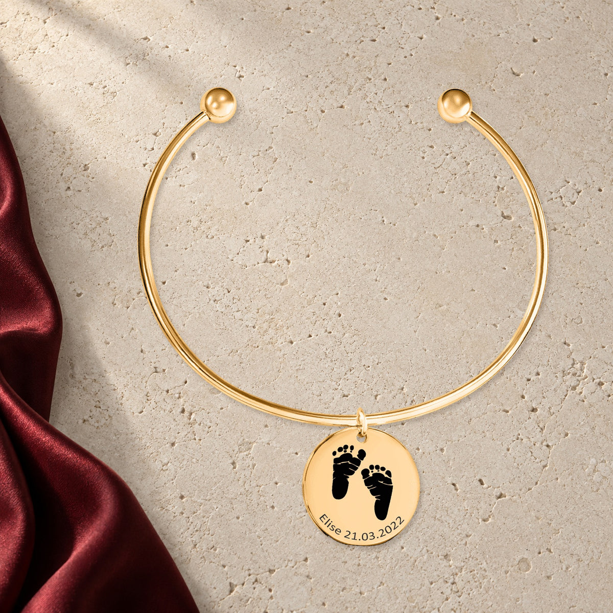 Bracelet jonc Empreinte de Pied Réelle Gravée en finition dorée sur pierre claire, avec étoffe rouge sanguine ; exemple de personnalisation.