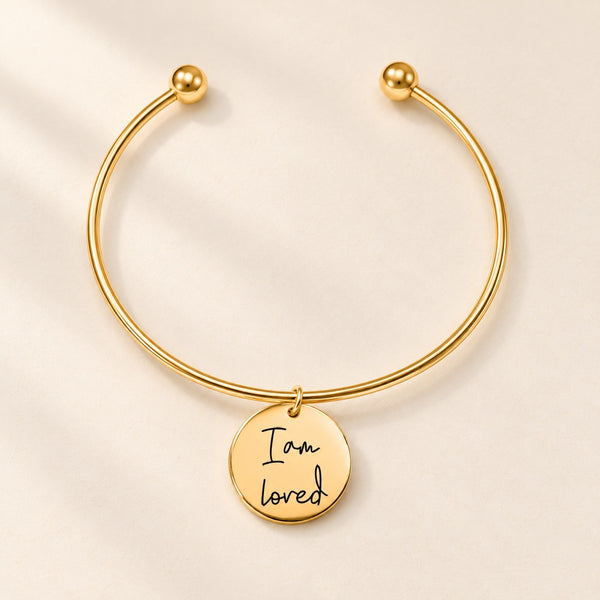 Bracelet jonc doré à médaille gravée « I am loved » sur fond ivoire.