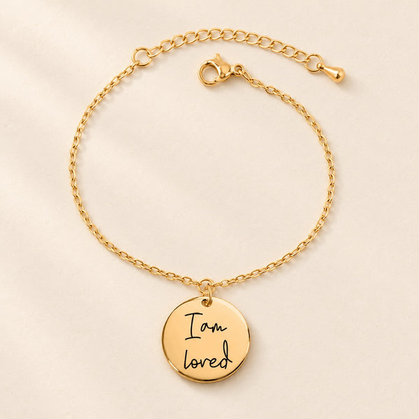 Bracelet chaîne doré à médaille gravée « I am loved » sur fond ivoire.
