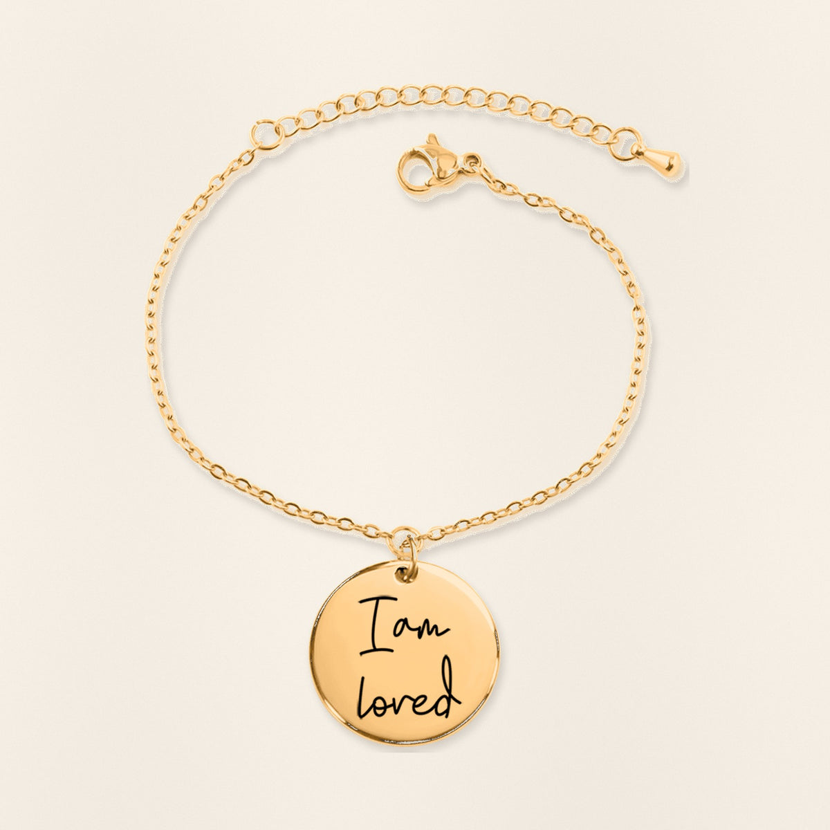 Bracelet Médaille Affirmations Personnalisée en finition dorée, exemple de gravure sur fond ivoire texturé.