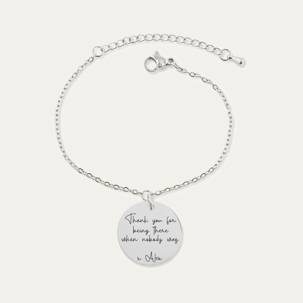 Bracelet chaîne en finition argentée, médaille gravée du message « Thank you for being there when nobody was — x Alex », sur fond ivoire.
