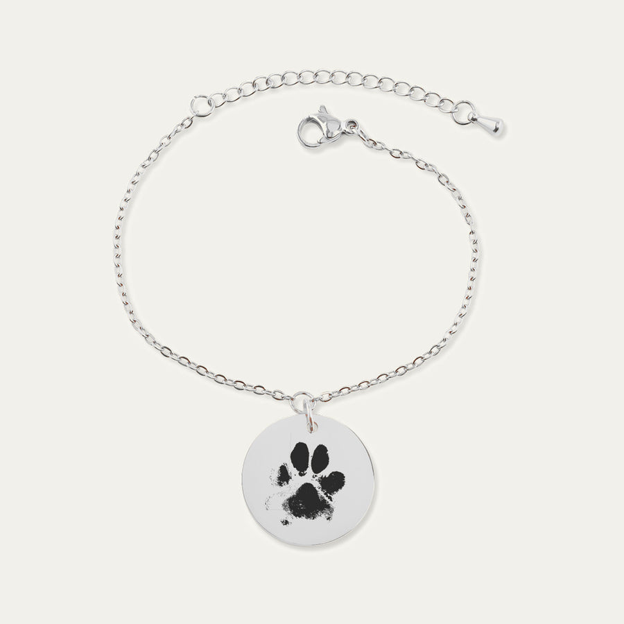 Bracelet chaîne en finition argentée, médaille gravée d’une empreinte de patte, sur fond ivoire.