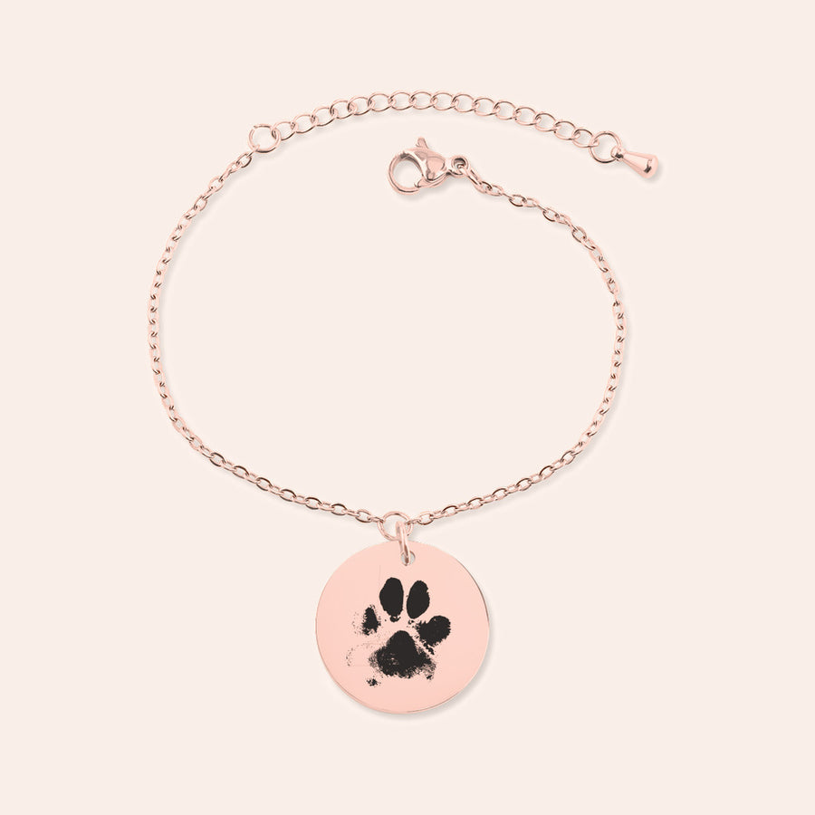 Bracelet chaîne en finition or rose, médaille gravée d’une empreinte de patte, sur fond ivoire.