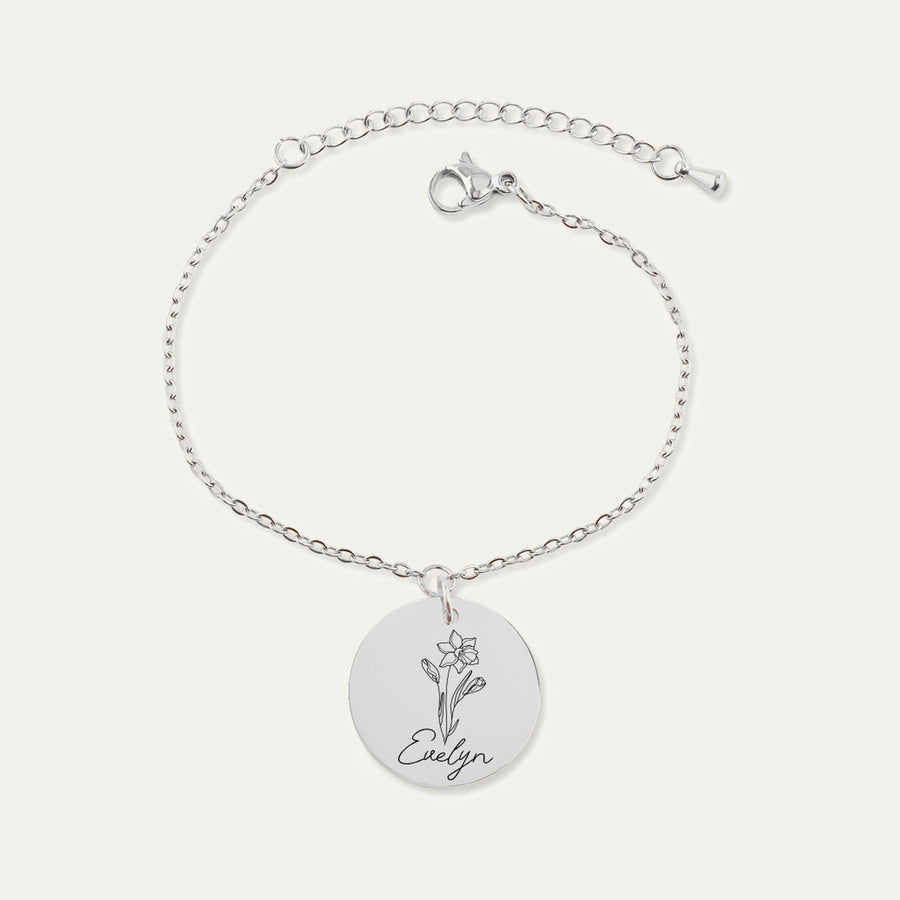 Bracelet chaîne en finition argentée, médaille gravée d’un narcisse et du prénom Evelyn, sur fond ivoire.