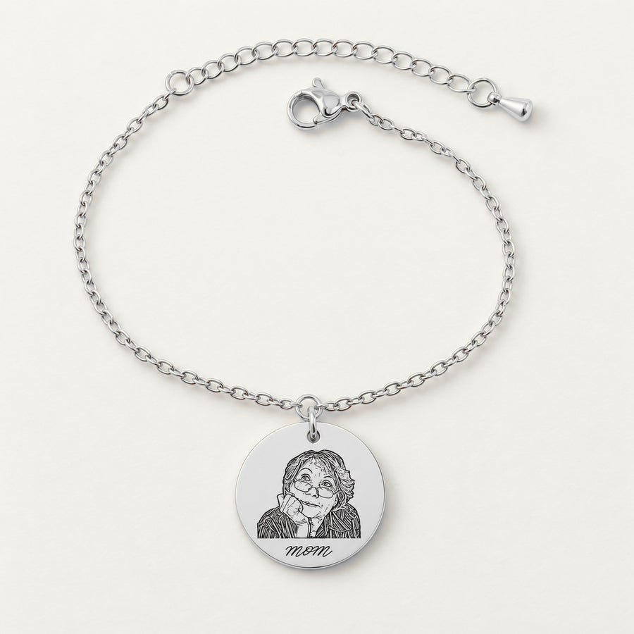 Bracelet chaîne en finition argentée, médaille gravée du portrait d’une femme avec lunettes et de la mention mom, sur fond ivoire.