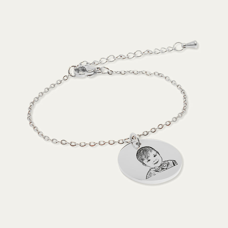 Bracelet chaîne en finition argentée, médaille gravée du portrait d’un bébé souriant avec un nœud papillon, sur fond ivoire.