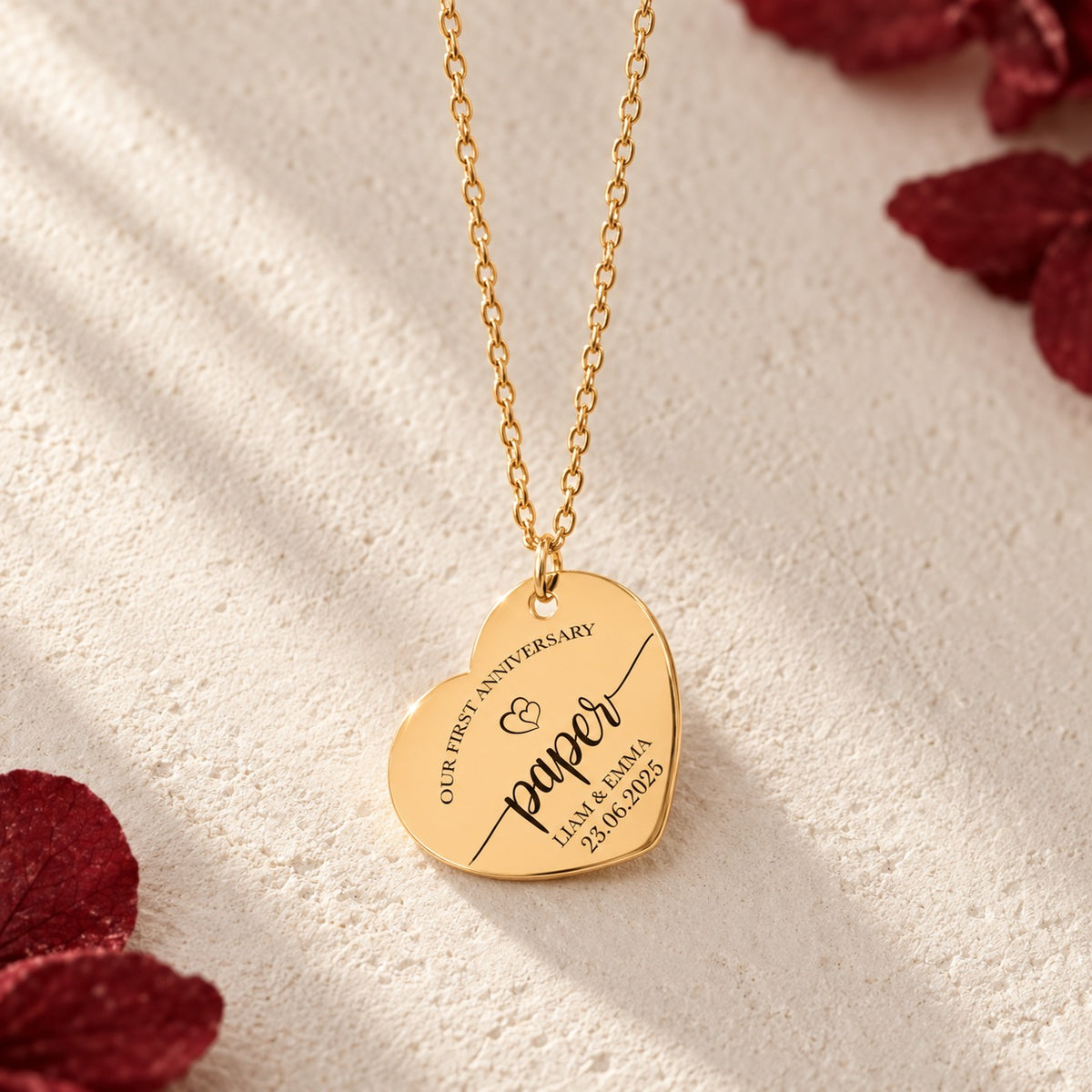 Détail du pendentif en forme de cœur doré avec inscription « paper », prénoms Liam et Emma et date, sur fond ivoire.