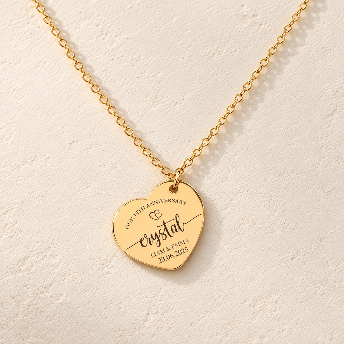 Collier Coeur Anniversaire de Mariage 15 ans doré, médaille en cœur gravée avec inscription « crystal », prénoms Liam et Emma et date 23.06.2025, sur fond ivoire.