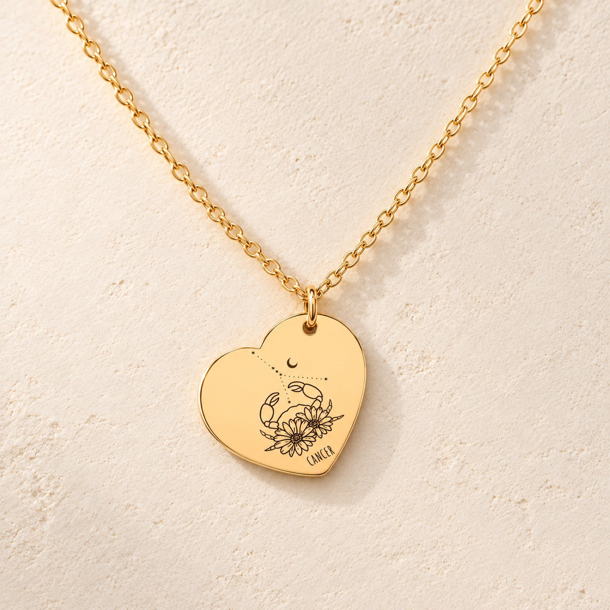 Collier cœur signe astrologique Cancer doré, médaille en cœur gravée avec motif du signe Cancer et ornements, sur fond ivoire.