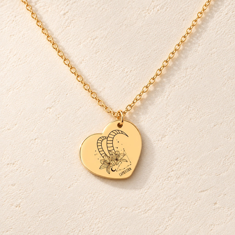 Collier cœur signe astrologique Capricorne doré, médaille en cœur gravée avec motif du signe Capricorne et ornements, sur fond ivoire.