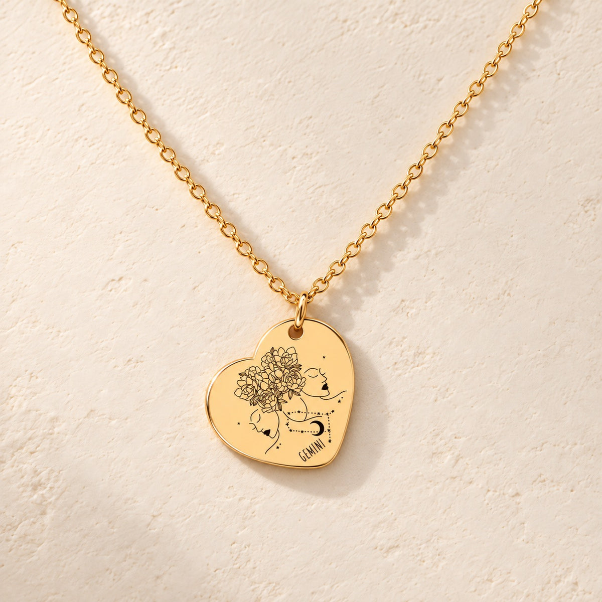 Collier cœur signe astrologique Gémeaux doré, médaille en cœur gravée avec motif du signe Gémeaux et ornements, sur fond ivoire.