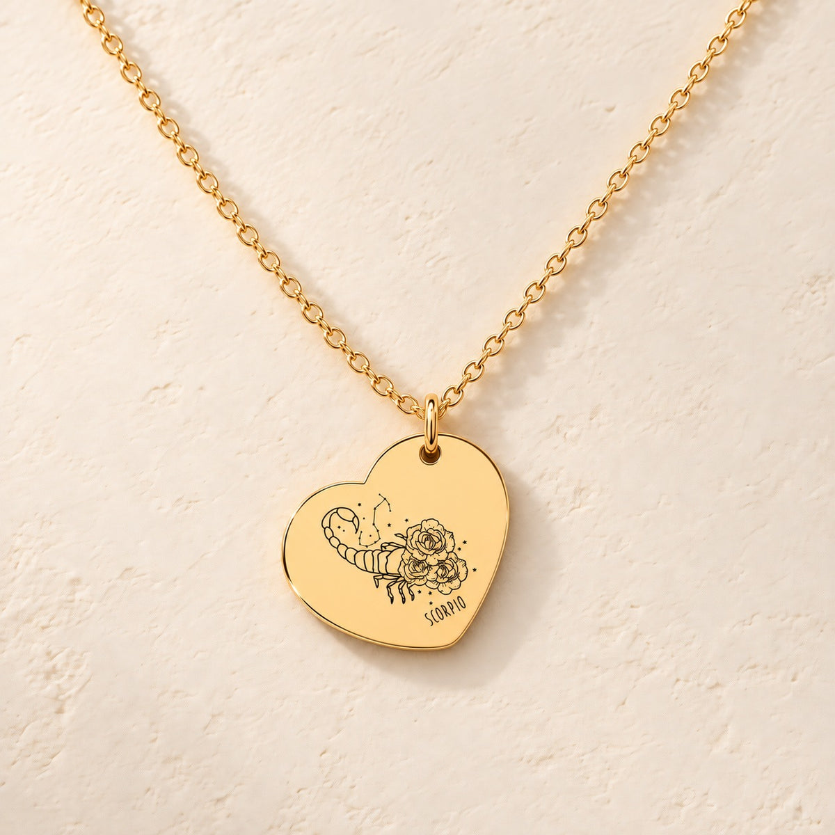 Collier cœur signe astrologique Scorpion doré, médaille en cœur gravée avec motif du signe Scorpion et ornements, sur fond ivoire.