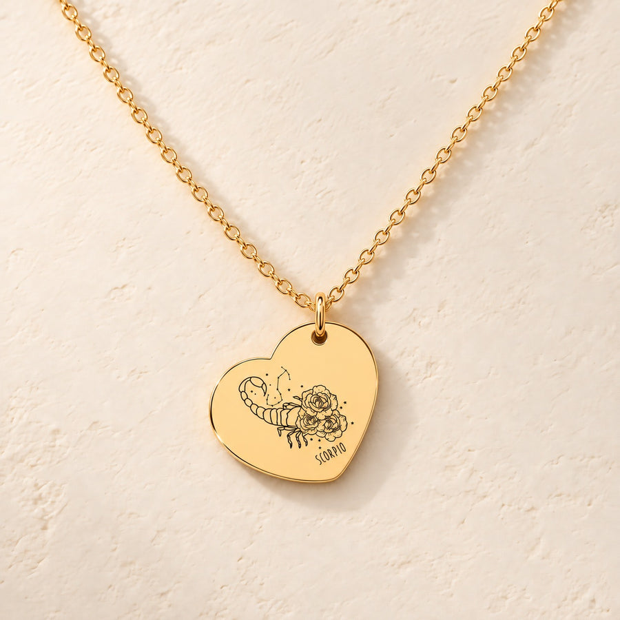 Collier cœur signe astrologique Scorpion doré, médaille en cœur gravée avec motif du signe Scorpion et ornements, sur fond ivoire.