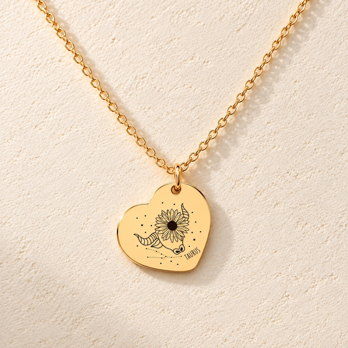 Collier cœur signe astrologique Taureau doré, médaille en cœur gravée avec motif du signe Taureau et ornements, sur fond ivoire.