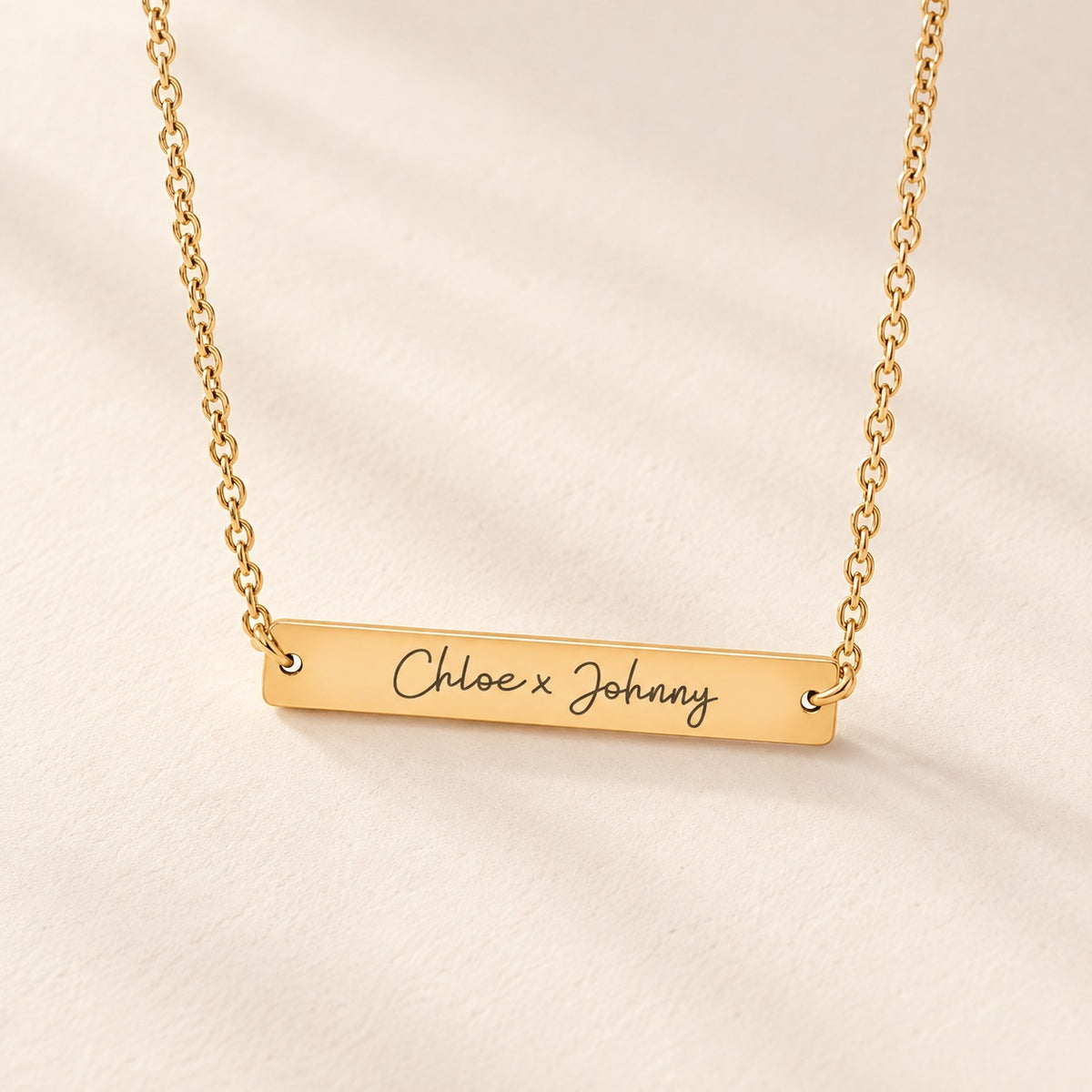 Collier Deux Prénoms doré, barrette gravée Chloe x Johnny sur fond ivoire.