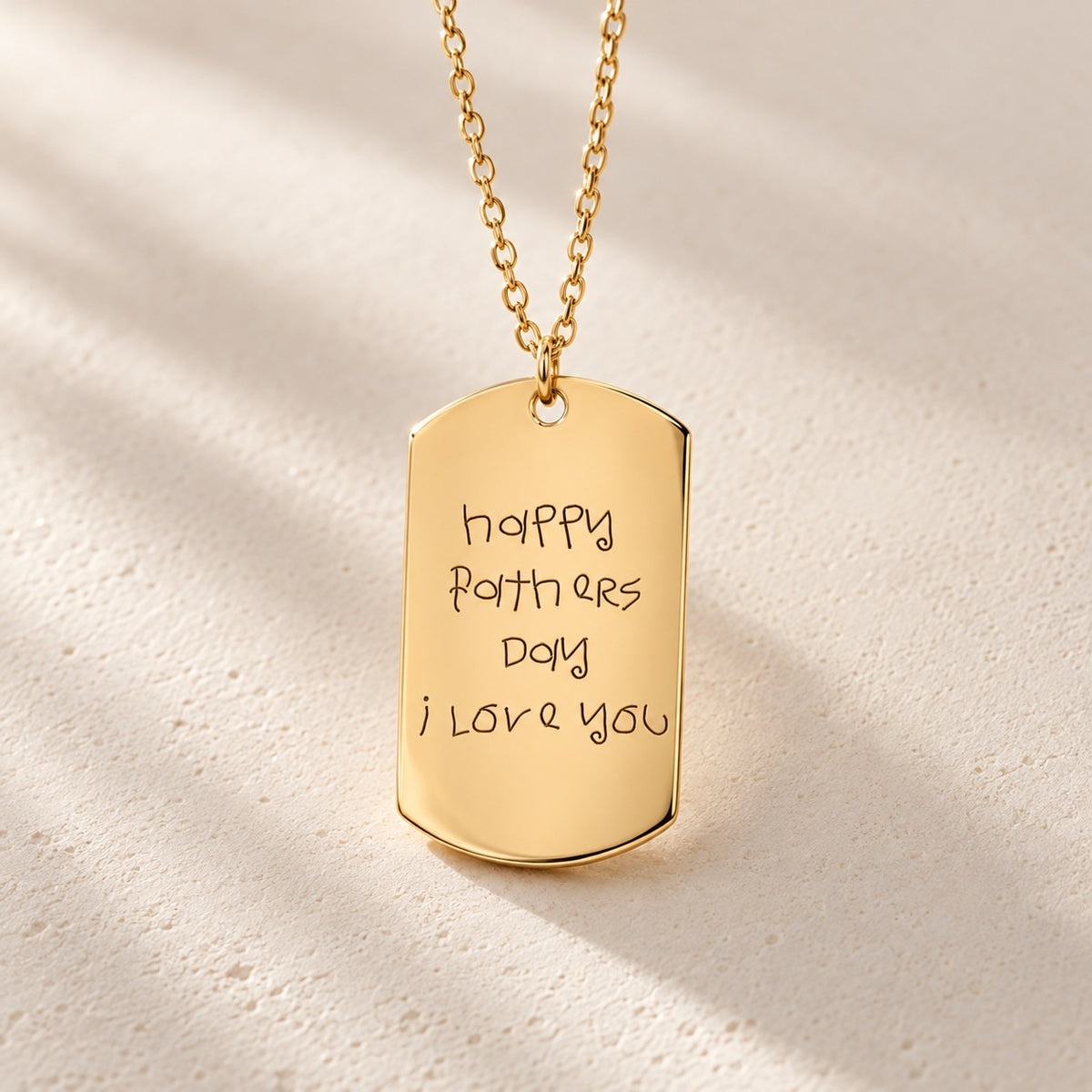 Détail du pendentif rectangulaire doré avec message d’enfant pour la fête des pères, sur fond ivoire.