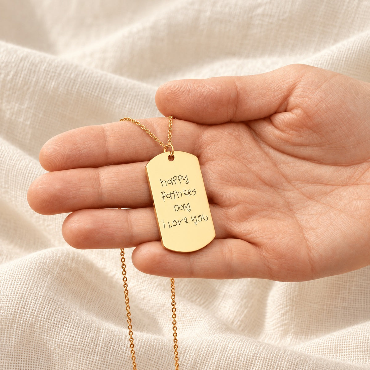 Collier Écriture Manuscrite doré présenté dans une main, avec pendentif rectangulaire gravé.