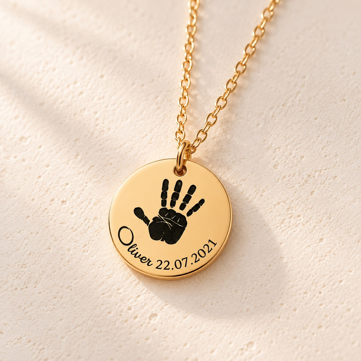 Détail de la médaille ronde doré avec empreinte de main, prénom Oliver et date 22.07.2021, sur fond ivoire.