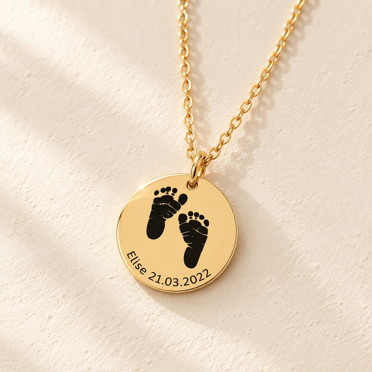 Détail de la médaille ronde doré avec deux empreintes de pieds, prénom Elise et date 21.03.2022, sur fond ivoire.