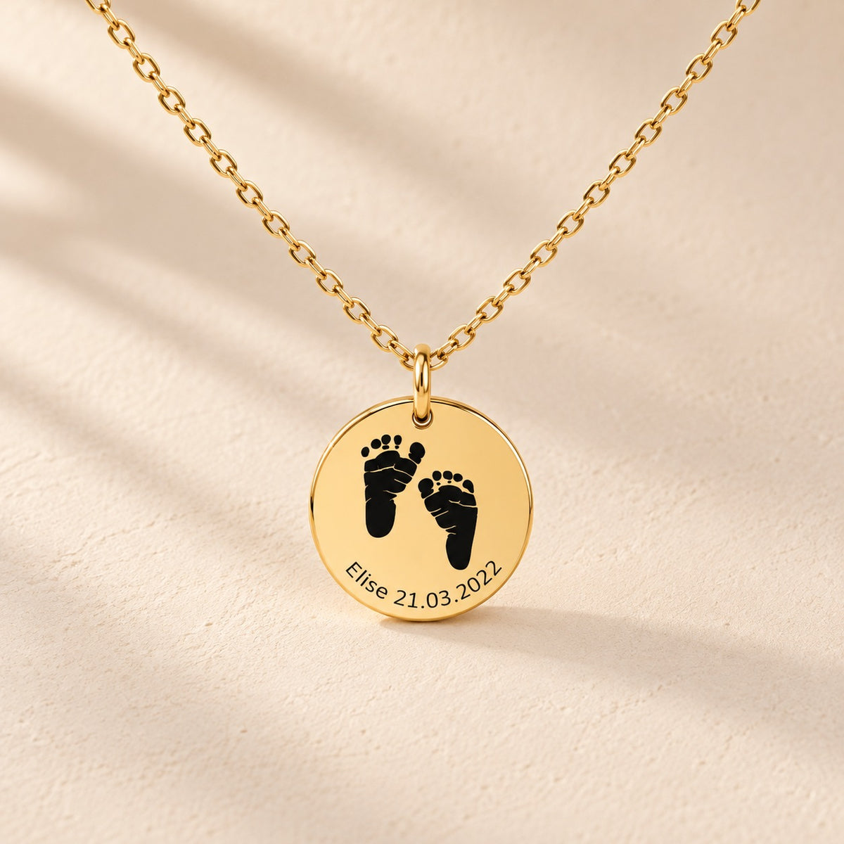 Collier Empreinte de Pied doré, médaille ronde avec deux empreintes de pieds, prénom Elise et date 21.03.2022 sur fond ivoire.