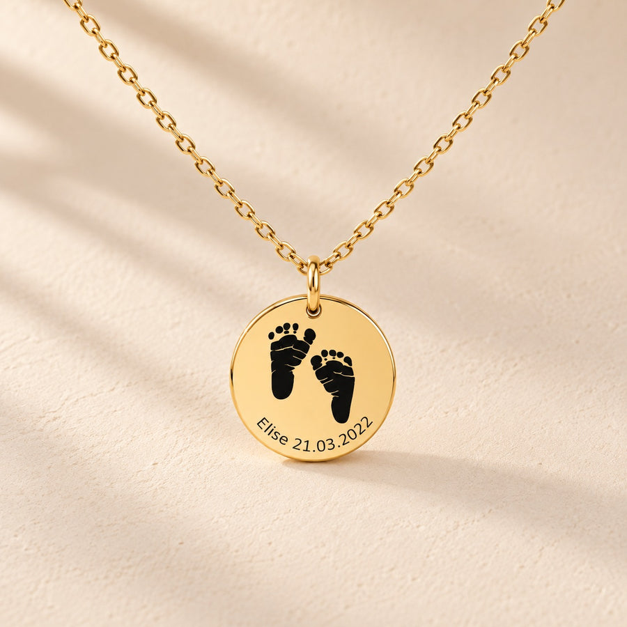 Collier Empreinte de Pied doré, médaille ronde avec deux empreintes de pieds, prénom Elise et date 21.03.2022 sur fond ivoire.