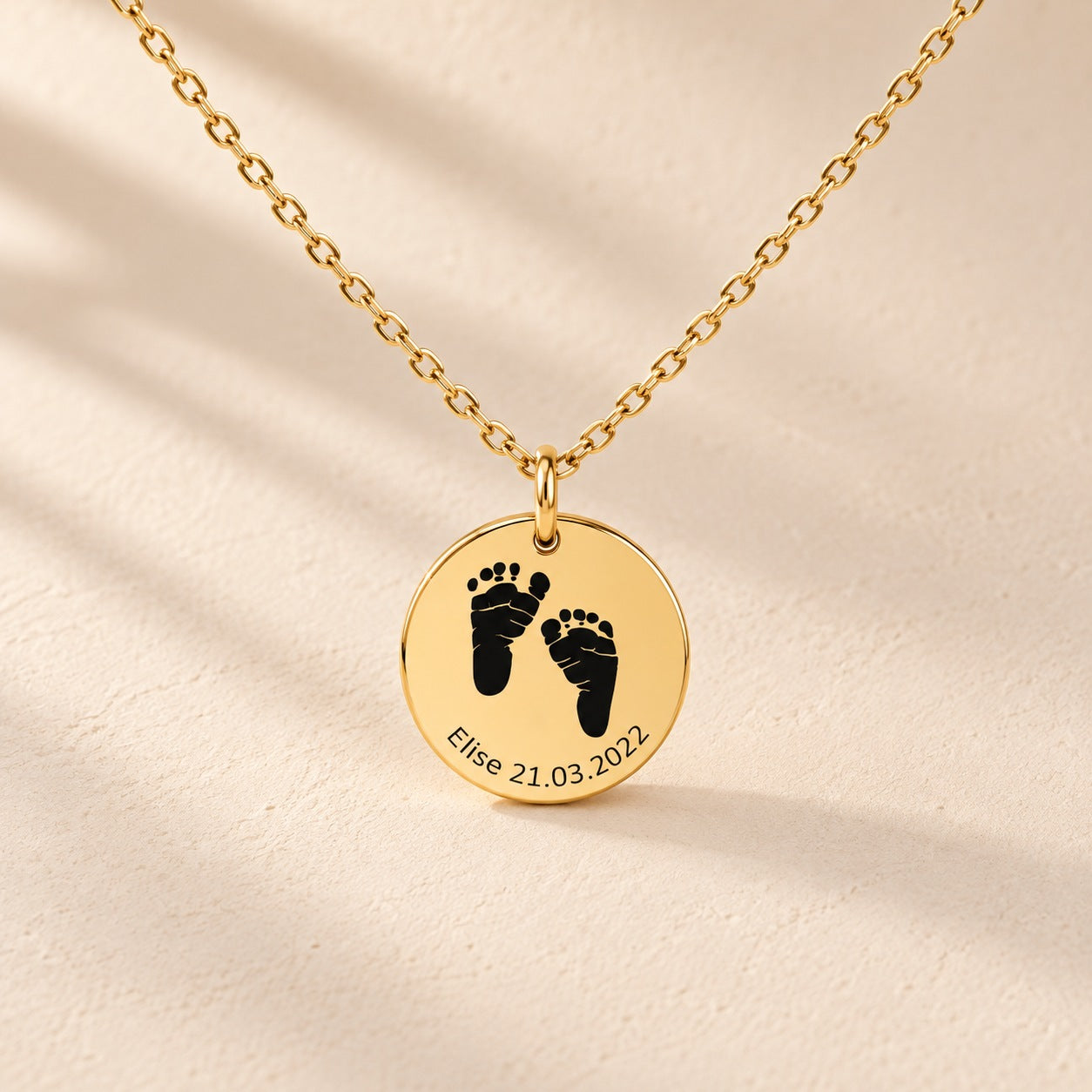 Collier Empreinte de Pied doré, médaille ronde avec deux empreintes de pieds, prénom Elise et date 21.03.2022 sur fond ivoire.