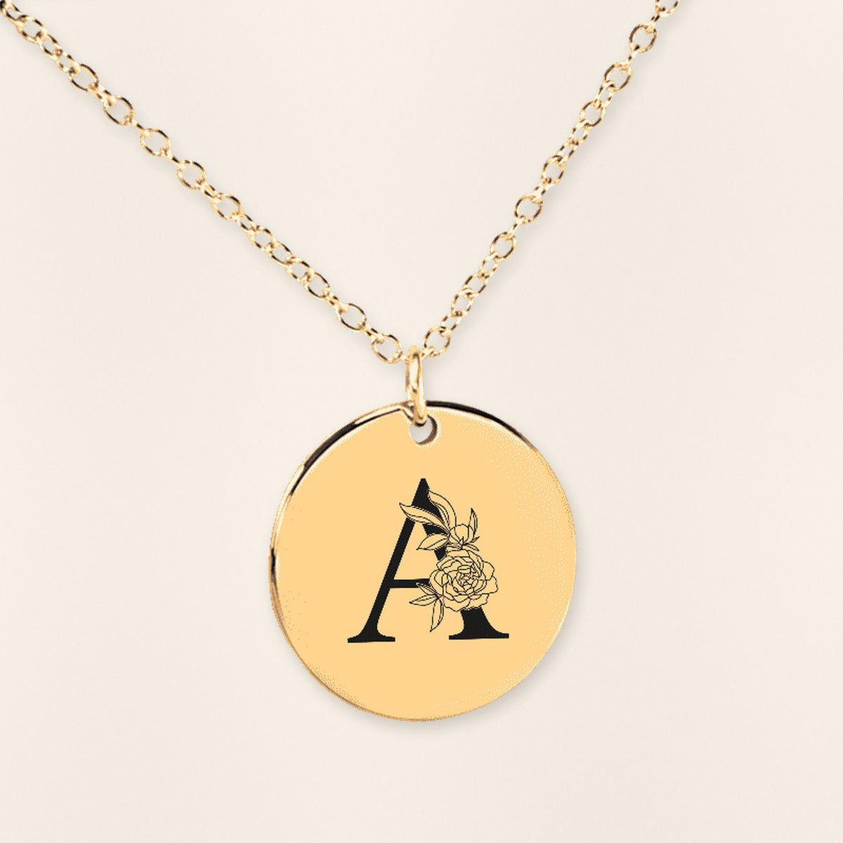 Collier doré à médaille ronde gravée de l’initiale A et de fleurs, sur fond ivoire texturé.