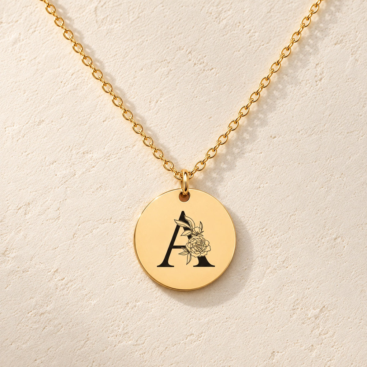 Collier initiale florale A personnalisé doré, médaille ronde gravée avec initiale A et fleurs, sur fond ivoire.