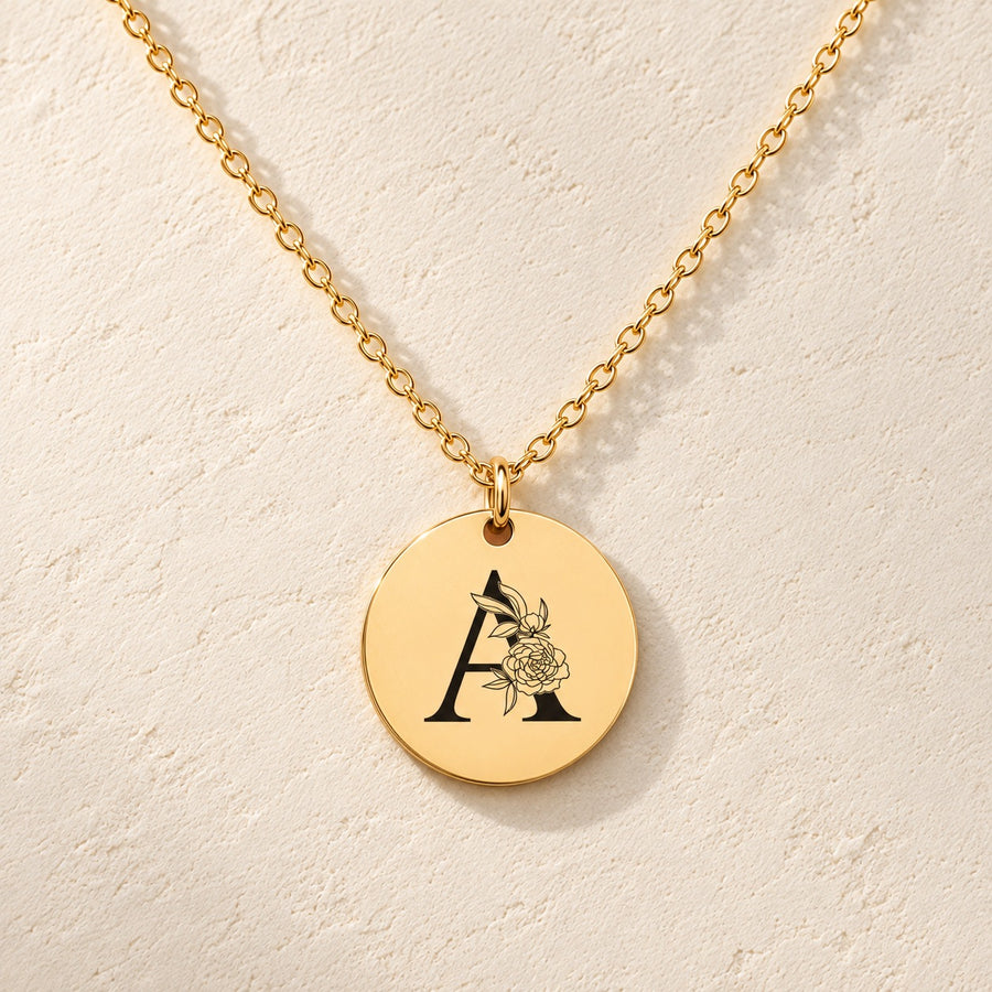 Collier initiale florale A personnalisé doré, médaille ronde gravée avec initiale A et fleurs, sur fond ivoire.