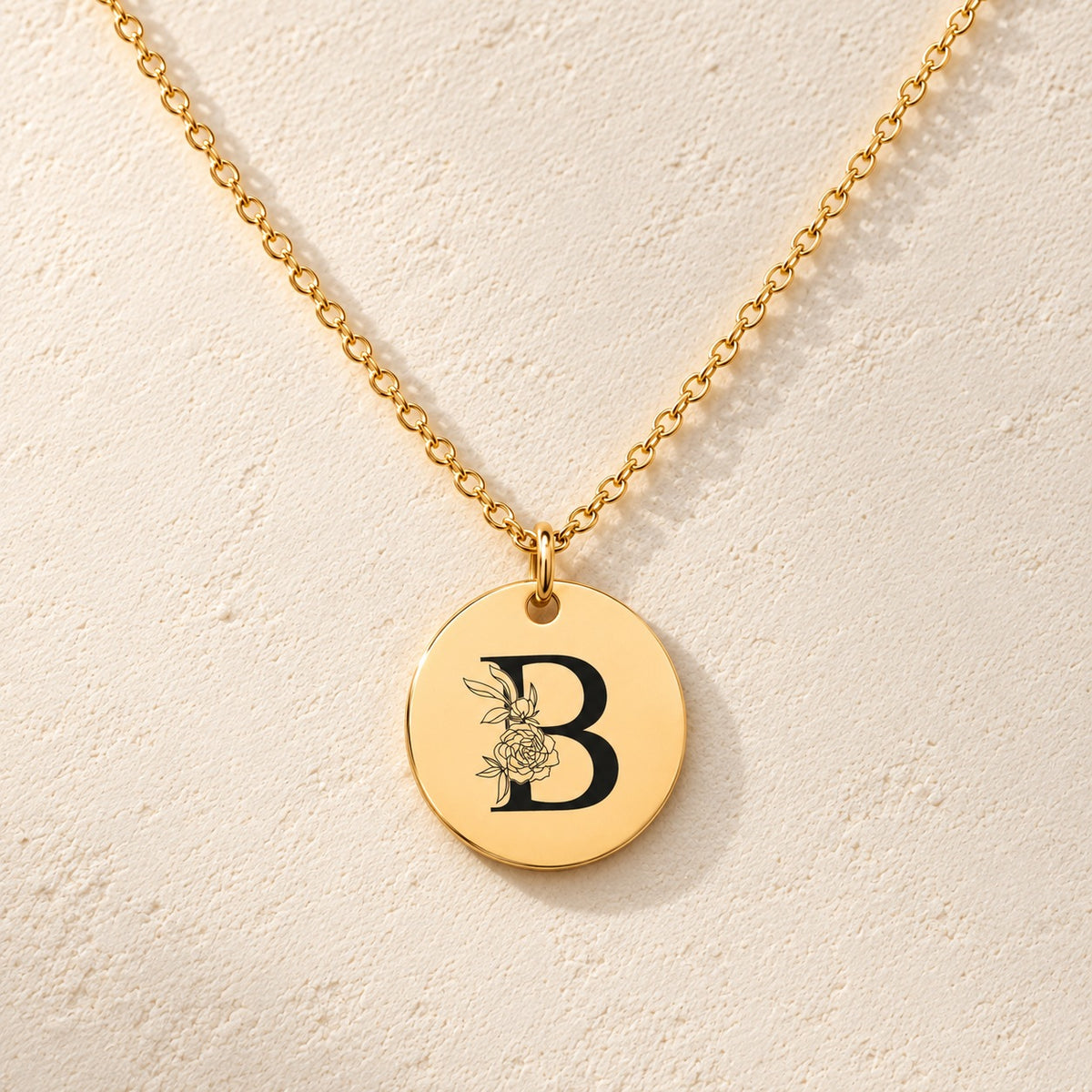 Collier initiale florale B personnalisé doré, médaille ronde gravée avec initiale B et fleurs, sur fond ivoire.