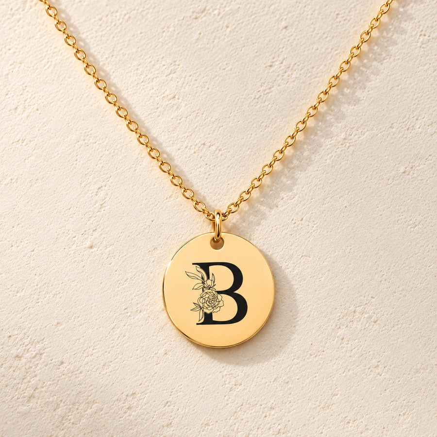 Collier initiale florale B personnalisé doré, médaille ronde gravée avec initiale B et fleurs, sur fond ivoire.