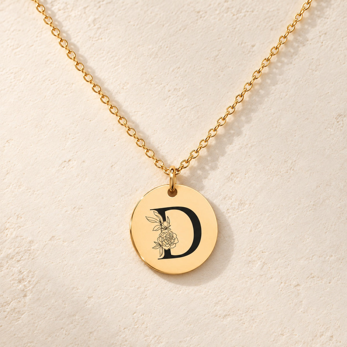 Collier initiale florale D personnalisé doré, médaille ronde gravée avec initiale D et fleurs, sur fond ivoire.