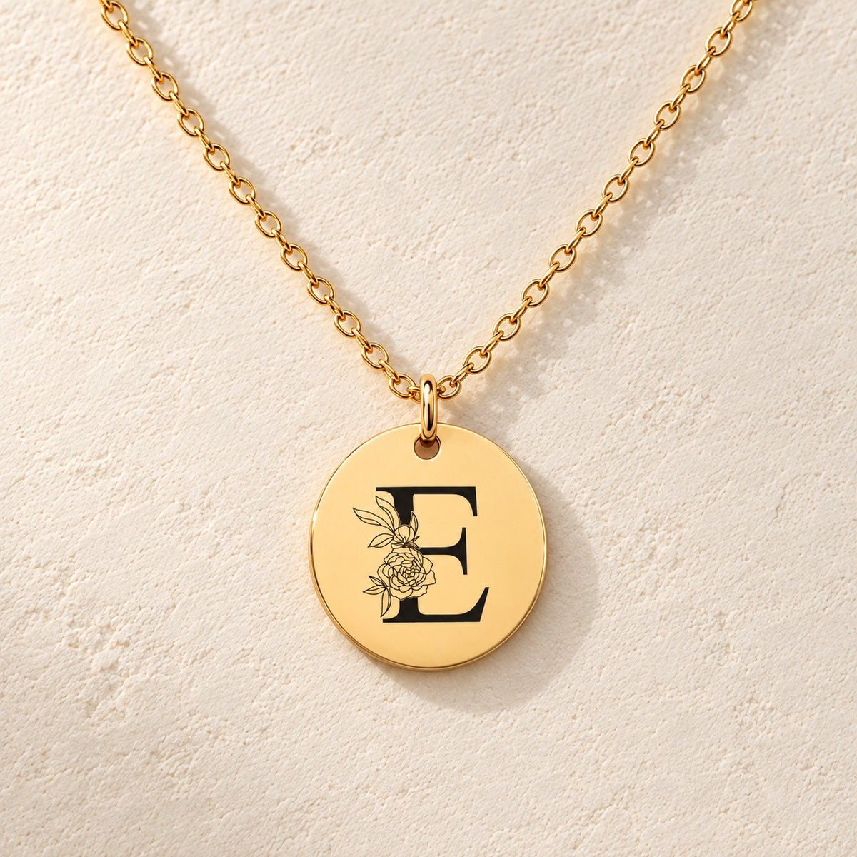 Collier initiale florale E personnalisé doré, médaille ronde gravée avec initiale E et fleurs, sur fond ivoire.