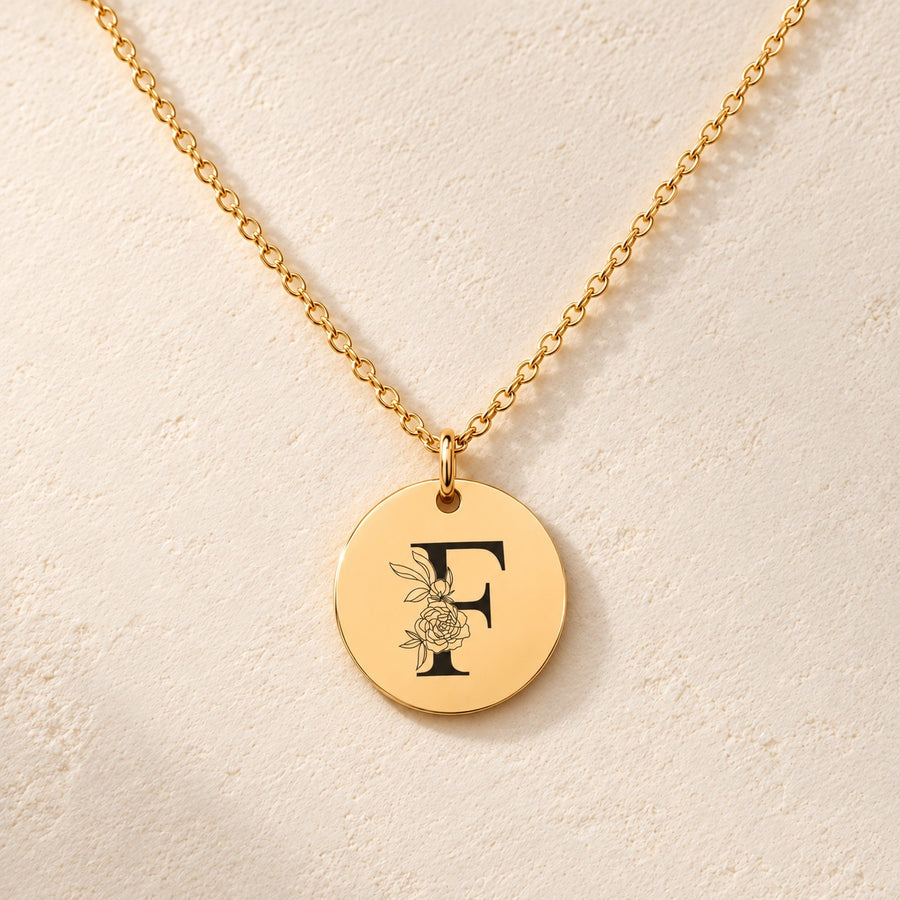 Collier initiale florale F personnalisé doré, médaille ronde gravée avec initiale F et fleurs, sur fond ivoire.