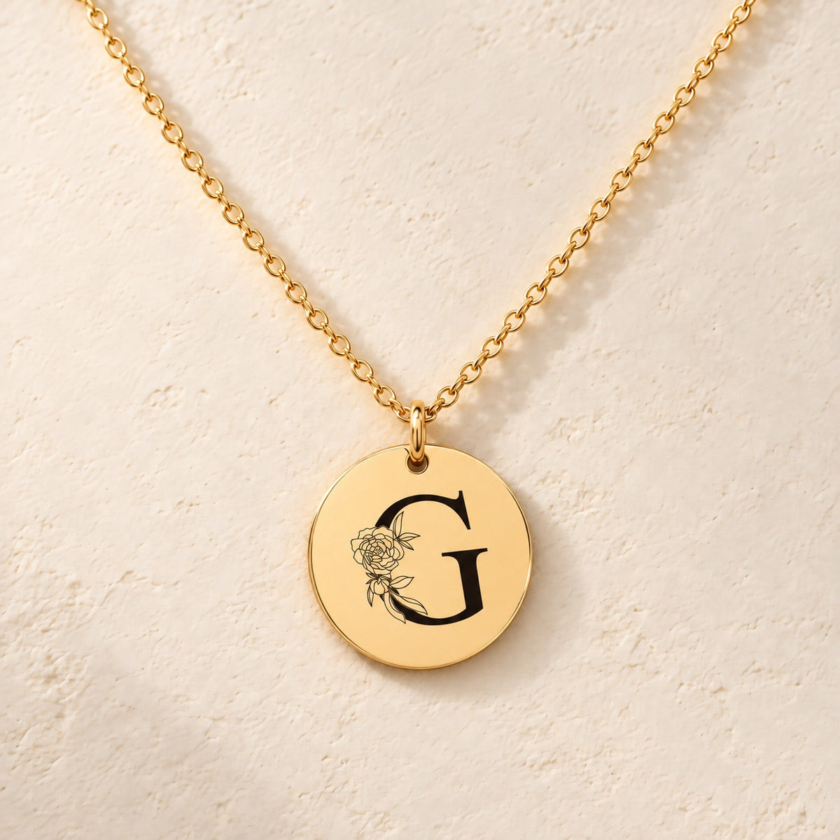 Collier initiale florale G personnalisé doré, médaille ronde gravée avec initiale G et fleurs, sur fond ivoire.