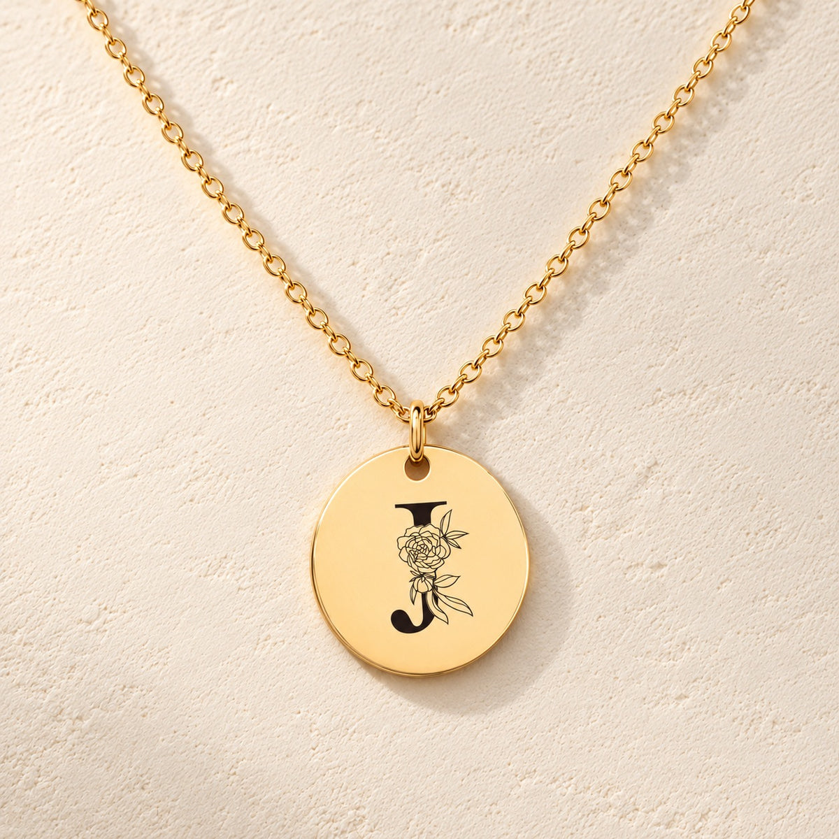 Collier initiale florale J personnalisé doré, médaille ronde gravée avec initiale J et fleurs, sur fond ivoire.