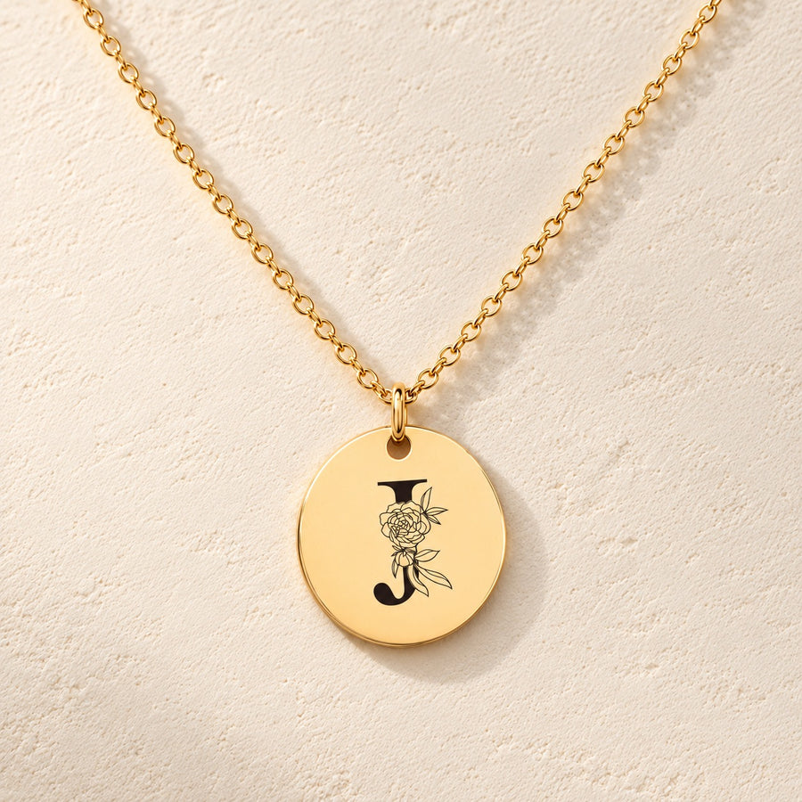 Collier initiale florale J personnalisé doré, médaille ronde gravée avec initiale J et fleurs, sur fond ivoire.
