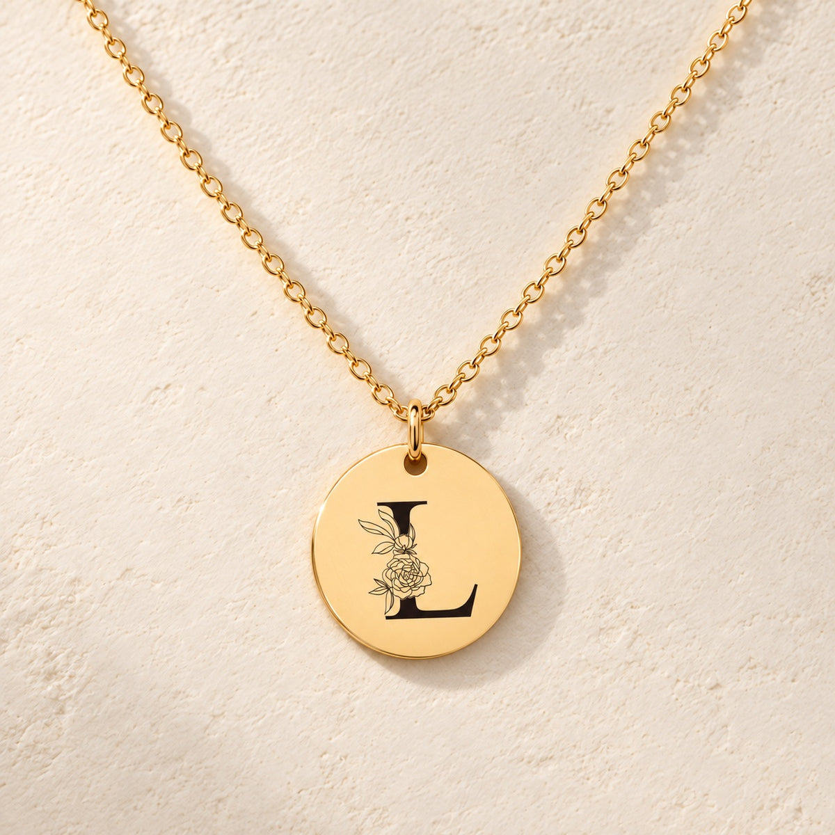 Collier initiale florale L personnalisé doré, médaille ronde gravée avec initiale L et fleurs, sur fond ivoire.