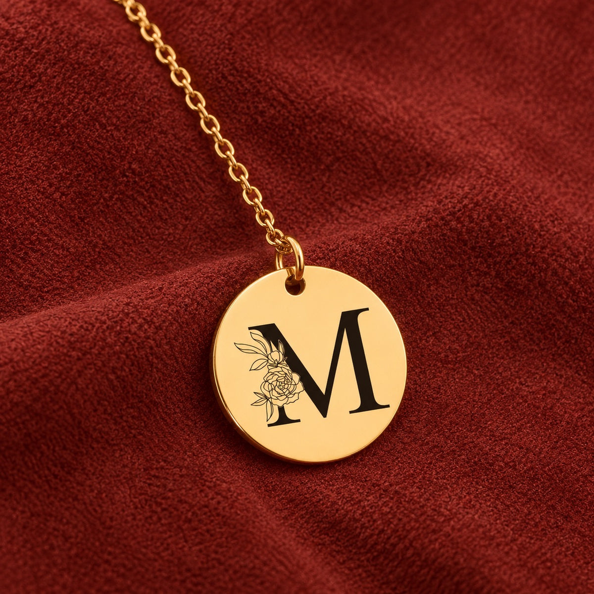 Médaille ronde dorée avec initiale M et fleurs, sur suédine rouge sanguine.