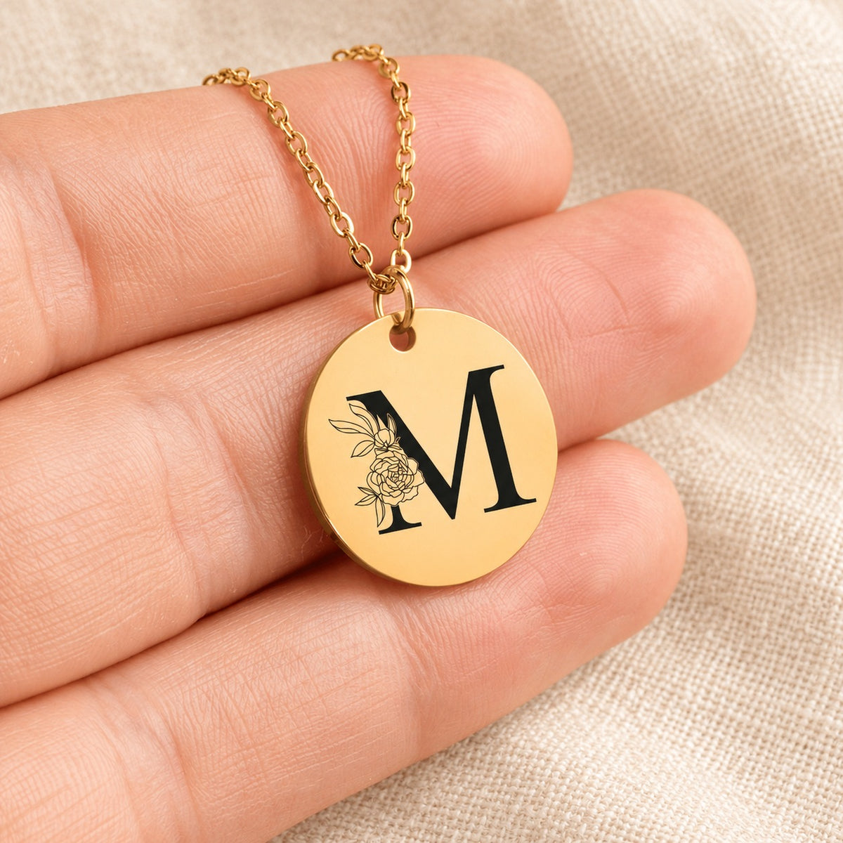 Collier initiale florale M personnalisé doré tenu en main, détail de la gravure.
