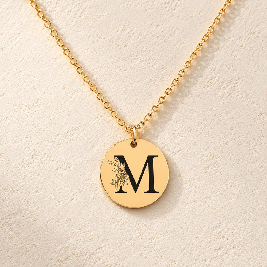 Collier initiale florale M personnalisé doré, médaille ronde gravée avec initiale M et fleurs, sur fond ivoire.