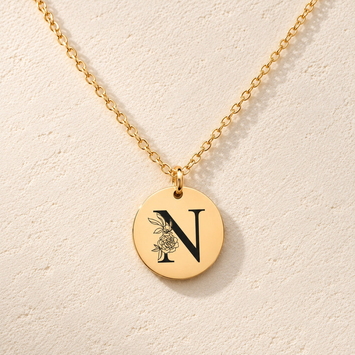 Collier initiale florale N personnalisé doré, médaille ronde gravée avec initiale N et fleurs, sur fond ivoire.