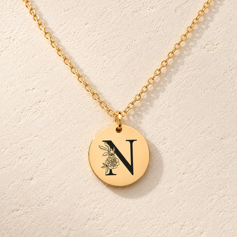 Collier initiale florale N personnalisé doré, médaille ronde gravée avec initiale N et fleurs, sur fond ivoire.