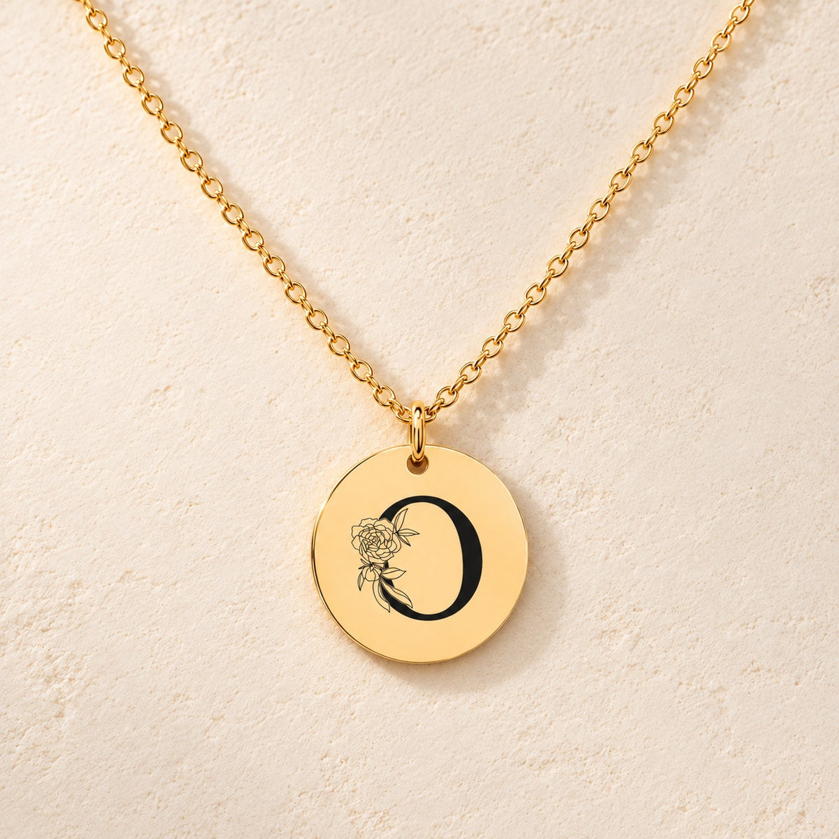 Collier initiale florale O personnalisé doré, médaille ronde gravée avec initiale O et fleurs, sur fond ivoire.